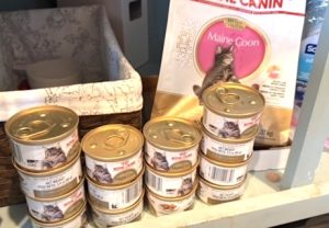 Piles of canned cat food sit on shelf next to bags of dry cat food.