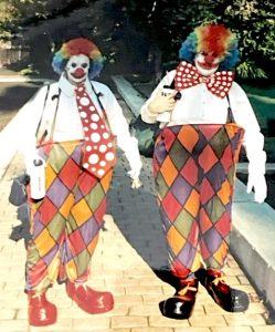A man and a woman are dressed up in mostly matching clown costumes on a paved park path with trees in the background. Both have white face paint and round clown noses, and are wearing rainbow afro wigs with white shirts. The woman on the left also wears an oversized red-and-white, polka-dotted necktie and red clown shoes, while the man on the right wears a similarly patterned bow tie and black clown shoes.