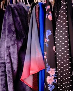 A close-up of many different blouses on hangers, including purple velvet, bright blue, floral, and polka-dotted.