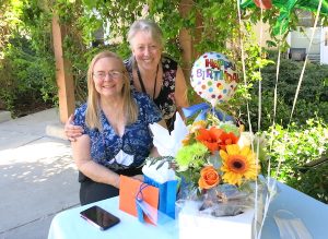 Two women, one seated and one standing, pose next to an outdoor table in a garden. On the table are a few wrapped gifts, a bouquet of bright flowers, and a mylar Happy Birthday balloon.
