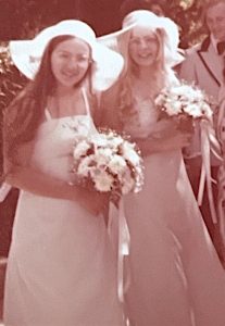 A vintage 1970s photo of two women in bridesmaid dresses and floppy sheer hats. Both women have long straight hair and glasses, one brunette and one blonde. 