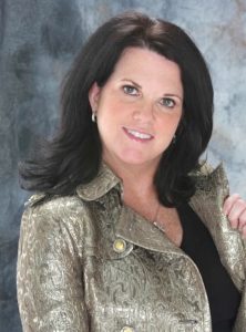 A brunette woman in a metallic brocade jacket with gold buttons poses for a glamour photo against a blue-grey backdrop.