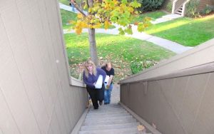 A man and a woman, each carrying packing supplies, climb a tall outdoor staircase. Green lawns, a leafy tree, and paved sidewalks can be seen on the condo grounds behind them.