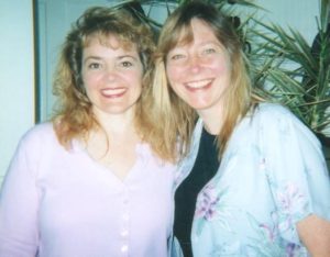 Two blonde women mile in an informal snapshot in front of some houseplants. The woman on the left wears a lavender button-down shirt, while the other woman wears a black t-shirt under a short-sleeved pale blue floral blouse.