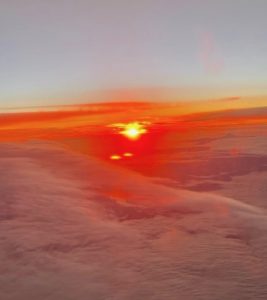 The view outside of an airplane window shows a burning orange and red sunrise glowing across the horizon, above layers of white clouds.