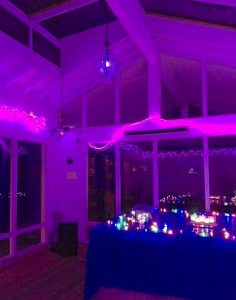 A glass walled atrium with very tall ceilings is lit up in a purple hue, while rows of bright multi-colored electric candles cover a nearby tabletop.