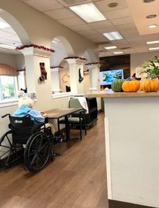A white-haired woman in a wheelchair is seated near the entrance of a dining room in a senior facility. On the left, a wall that separates the space features wide arches so you can see out windows across the room. Pumpkins are in a seasonal display on a counter in the foreground.