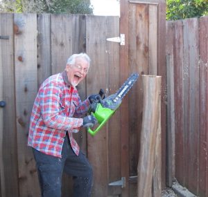 A laughing, silver-haired man poses with a chainsaw in a backyard. He is wearing jeans, a heavy plaid shirt, and glasses.