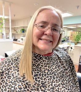 A snapshot of a blonde woman in her fifties, smiling while she gets a haircut in a small salon. She is wearing glasses as well as a leopard-print salon cape on her shoulders.
