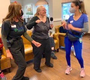 In a living room setting, a blonde woman (wearing glasses and a grey t-shirt bearing an illustration of a cat), is singing into a karaoke microphone. Dancing alongside her are two young women staffers, wearing medical masks and name tags.