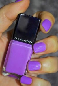 A bottle of opaque purple nail polish is held in the hand of a woman whose nails are painted with that same purple polish.