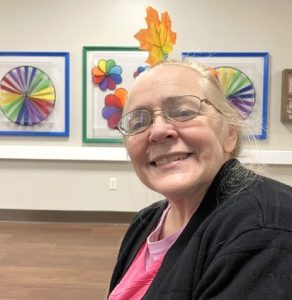 A smiling blonde women, wearing a pink shirt and a black cardigan sweater, poses with a leaf propped up on the top of her head. In the background are framed rainbow artworks.