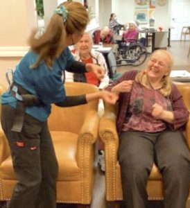A smiling blonde woman is seen dancing in her chair, while a staff member stands near her, also dancing. Other senior care residents can be seen seated in the background.