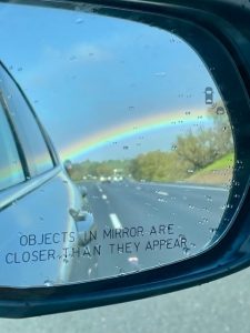 A car's rear-view side mirror shows a beautiful rainbow that stretches across a blue-grey sky. 