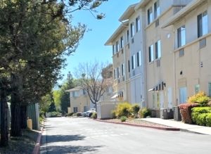 A view of a long, wide, paved road behind a large senior residence building. There are trees on the left, and the building on the right. Outside the building, in the foreground, are bushes and other landscaping. In the far background can be seen adjacent buildings and some parked cars.