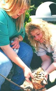 Two blonde women, one seated and one crouching, pet a young wild cat on a leash.
