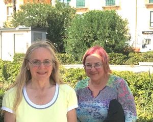 A blonde woman in a yellow t-shirt poses in a small town in Sicily next to a woman with purple hair in a brightly-patterned shirt. Both women are wearing wire-framed glasses. Behind the women can be seen green bushes and a large building housing a restaurant.