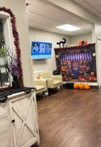 A small lobby area in a memory care unit features armchairs and a flatscreen TV on the wall. In the background is a large Halloween-themed wall hanging and decorative pumpkins.
