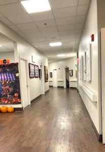 A hallway in a memory care unit features cream-colored walls, plank flooring, lots of doors to residents' rooms, and framed artwork on the walls.