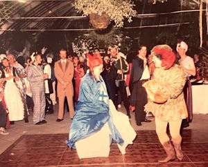 In a vintage color snapshot, costumed wedding guests surround a wooden dance floor, where the costumed bride and groom begin their first dance.