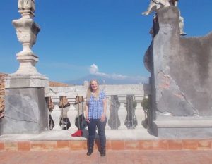 A blonde woman in a blue patterned blouse and glasses poses in front of Sicily's Mount Etna. The volcano far in the background against a blue sky. 