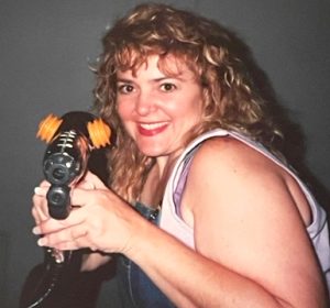 A blonde woman smiles for the camera while holding a silver futuristic ray gun. 