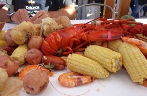 On a buffet table at the Deloach winery is spread an assortment of cooked items, including a bright red lobster, garlic, new potatoes, corn on the cob, prawns, and sausages.