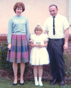 A vintage snapshot from the 1960s shows a brunette woman in a blue and fuchsia plaid skirt and blue blouse, a man in a white shirt with a black necktie and and black dress pants, and a young blonde girl dressed in white for her first holy communion. They are posed outside on a lawn. 