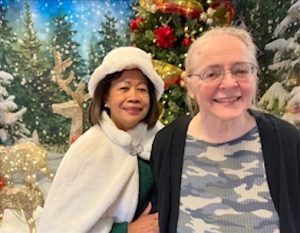 A brunette woman, wearing a white furry hat and holiday cape, poses with a blonde woman with glasses in front of a winter holiday scene. The blonde woman is wearing a blue, grey, and white camouflage-patterned shirt and black sweater. Both women are smiling for the camera.