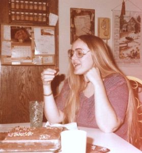 A vintage, sepia-toned snapshot from the 1970s shows a woman with very long, straight blonde pair and glasses about to pop a piece of chocolate cake into her mouth. On the table in front of her is the rest of the birthday cake, frosted in chocolate.