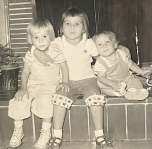 A vintage black-and-white snapshot shows three young children sitting in a row on the brick hearth of a fireplace. From left to right are a blonde girl, a slightly older brunette girl, and a little blonde boy younger than both girls. The older girl in the center has her arms around both siblings.