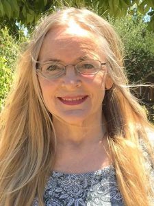 A smiling blonde woman wears wire-framed glasses and a paisley black-and-white shirt. Behind her are shown various green trees and bushes.