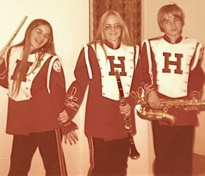 A brunette teenage girl in glasses, a blonde teenage girl in glasses, and a blonde teenage boy pose in their high school band uniforms. They wear red jackets with white fronts and shiny buttons and black pants with red stripes down the outer legs. Each teenager holds their band instrument.