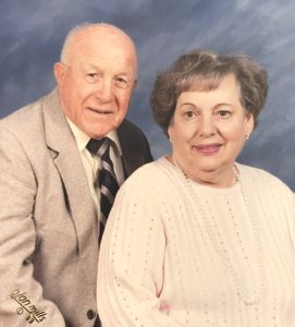 A 1999 professional portrait photo shows a senior couple smiling softly against a blue backdrop. On the left, the man wears a grey sport coat, white dress shirt, and striped tie. On the right, his wife is wearing a cream-colored sweater, a string of pearls, and pearl earrings.