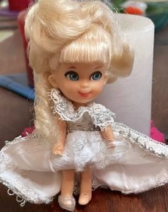 A 3-inch-high, collectible vinyl doll known as a Liddle Kiddle is dressed as Cinderella. She has a head full of fancy blonde hair, a white princess dress trimmed in silver, and transparent shoes. Her painted, childlike facial features include pink lips and oversized blue eyes. 