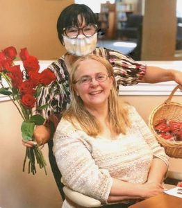 Two smiling women with glasses pose for this snapshot; one is a seated blonde woman in a cream-colored sweater, while an Asian woman with short black hair stands behind her. The standing woman holds a basket in her left hand, and a bouquet of red roses in her right. 