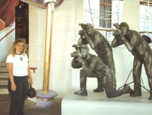 A smiling blonde woman in dark pants and a white t-shirt cleverly poses in front of three bronze statues of celebrity photographers. 