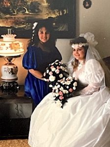 In this vintage snapshot, a smiling bride perches on a chair in a bright white wedding gown with voluminous skirt, plus a headband veil. To her right is her brunette maid of honor in a royal blue satin gown. Both women hold large flower bouquets.