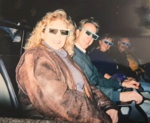 A blonde woman in a brown leather jacket and 3-D glasses smiles at the camera from her movie theater seat. Beside her is seated a man, woman, and another man; all are smiling at the camera and wearing 3-D glasses.