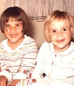 In this vintage snapshot, a brunette little girl and a blonde little girl with matching bob haircuts smile mischievously at the camera.