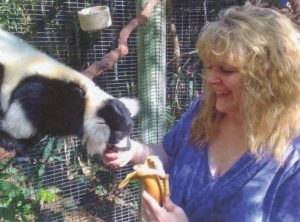 A smiling blonde woman in a blue-purple shirt feed a banana to a black-and-white ruffled lemur. 