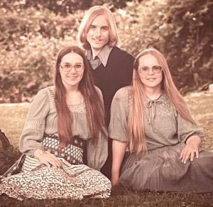In this vintage formal portrait, three smiling siblings in their 20s pose seated on grass next to the base of a large tree. In front is a brunette woman and a blonde woman, both with long hair and eyeglasses. The women are wearing soft, light-colored blouses and skirts. Behind them kneels a blonde man wearing a dark v-necked sweater over a dress shirt.