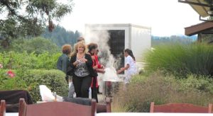 A blonde woman walks happily towards an outdoor buffet table at a winery, after checking on the progress of lobster being cooked for the crowd. Behind her can be seen a food serving area, above which lofts a cloud of steam.