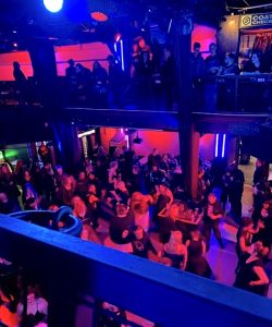A large, mixed crowd of people, most of them clad in black, can be seen dancing on the main dance floor and hangin out on the second-story, wraparound balcony of the DNA Lounge night club.