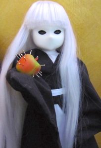 An extremely stylized, spooky, goth-inspired collectible doll features a mask-like white face with almond-shaped blank black eyes and a faint sculptured bump for a nose. She has long white hair with bangs, and is dressed in a black kimono-like robe with white sash. She is holding a tiny red-and-green apple with nails sticking out of it.