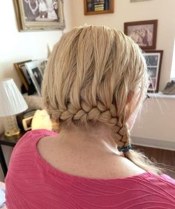 This snapshot of the back of a blonde woman's hair shows an elaborate braid that curves around the base of her head. The woman is wearing a textured pink shirt and sitting on a bed; on her nightstand rests a lamp and a glowing, cat-shaped nightlight. In the background, framed photos are on the walls, dresser, and windowsill.