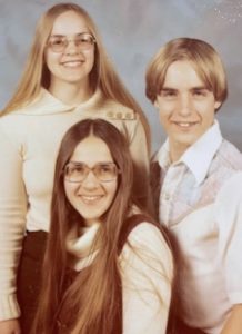 A blonde young woman, a brunette young woman, and blonde young man pose for a formal sibling portrait circa 1976 or 1977. The women are wearing glasses and have long straight hair parted in the center.