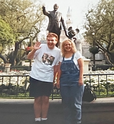 A blonde man and blonde woman pose in front of the statue of Walt Disney and Mickey Mouse on the grounds of Disneyland. Cinderella's castle can be seen behind them in the distance. Both are in white t-shirts; the man also wears black shorts and the woman wears denim overalls.