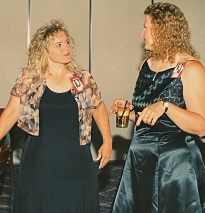 Two blonde women chat in a hotel ballroom. Both are wearing dresses and class reunion name tags.
