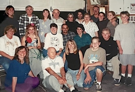 A large group of more than 20 family members and friends, dressed casually, gather in a suburban garage for a group snapshot. There are men and women of various ages pictured alongside some older kids.